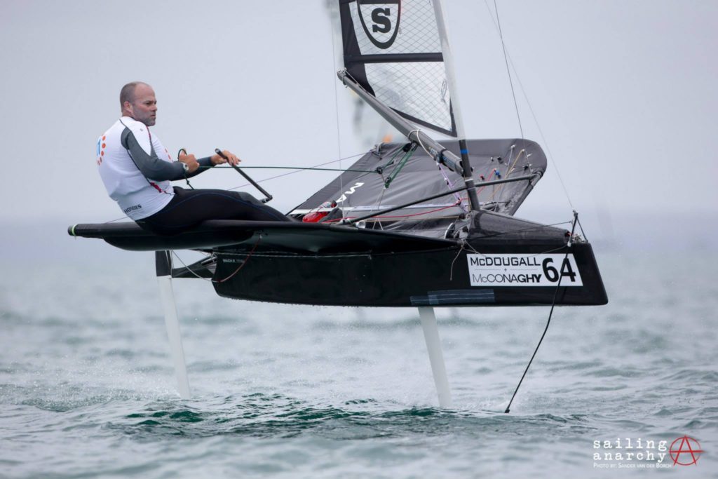 A man sails a black hydrofoil boat labeled “64” above the water’s surface, wearing a white shirt and black pants. The sea is calm, and the boat appears to be gliding swiftly. “Sailing Anarchy” is visible in the corner.