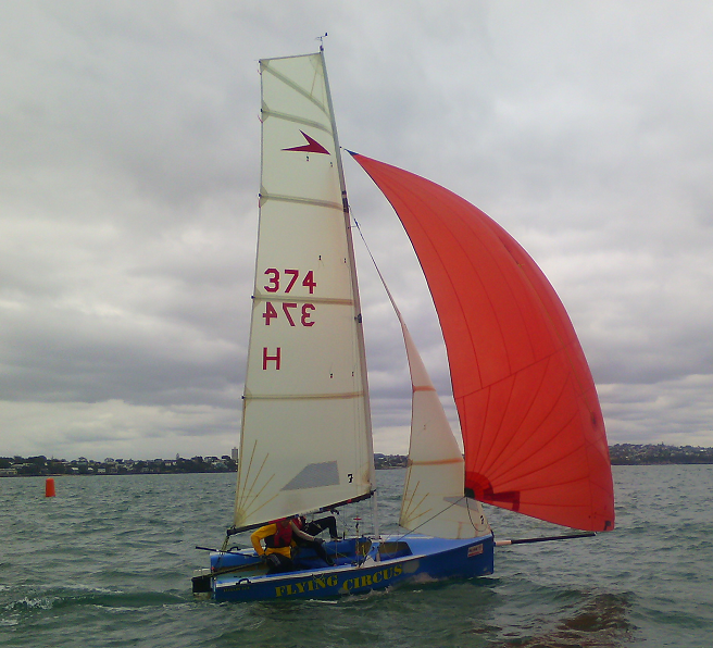 A small blue sailing boat named 'Flying Circus' with two people aboard and a large red sail glides across the water under a cloudy sky; shoreline and buildings are visible in the background.