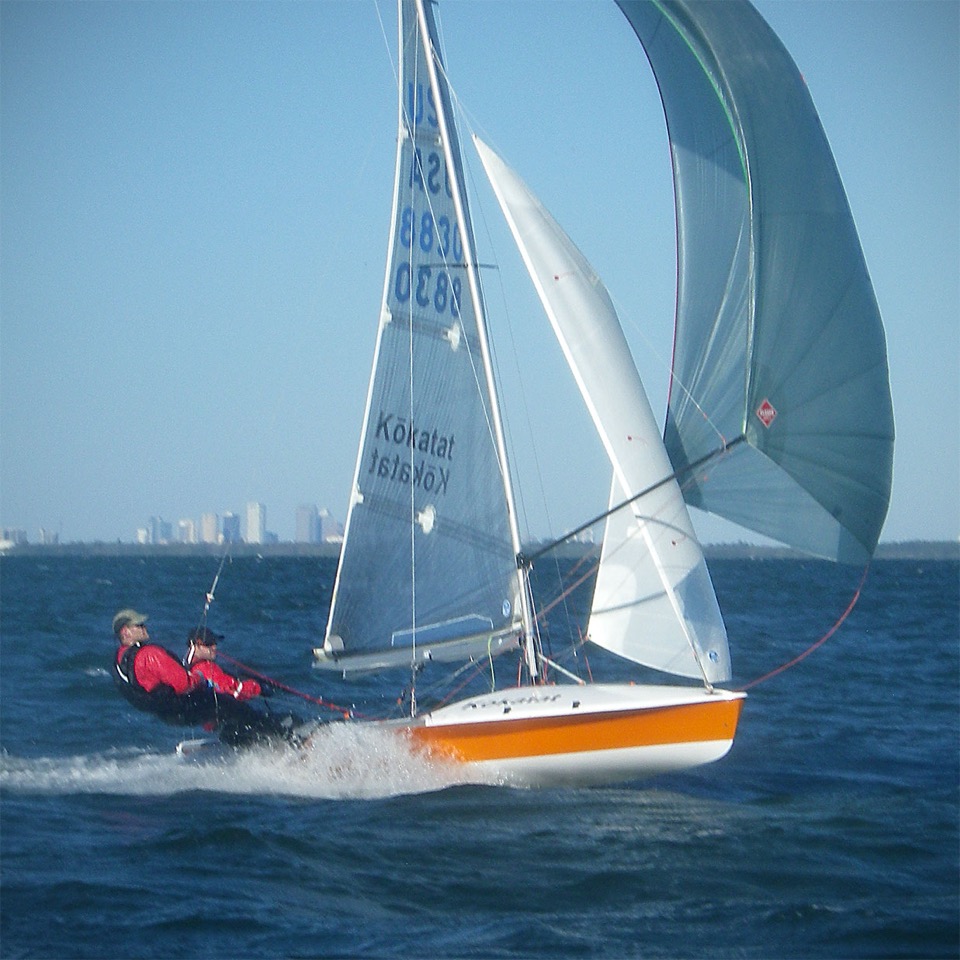 Two sailors in red jackets are sailing a small yellow and white sailboat at high speed on open water, leaning back with harnesses, with city buildings visible in the distant background.