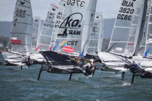 Several sailboats with hydrofoils race closely together on choppy water, their sails marked with country codes and numbers. Sailors lean out over the sides, and mountains are visible in the background.