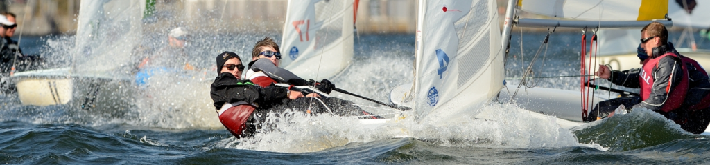 A group of people in life jackets and sunglasses sail small boats at high speed, creating splashes and waves as they compete in a regatta on a sunny day.