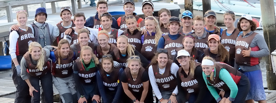 A large group of smiling young adults wearing matching brown 'Brown' vests pose together on a dock with boats and water in the background.
