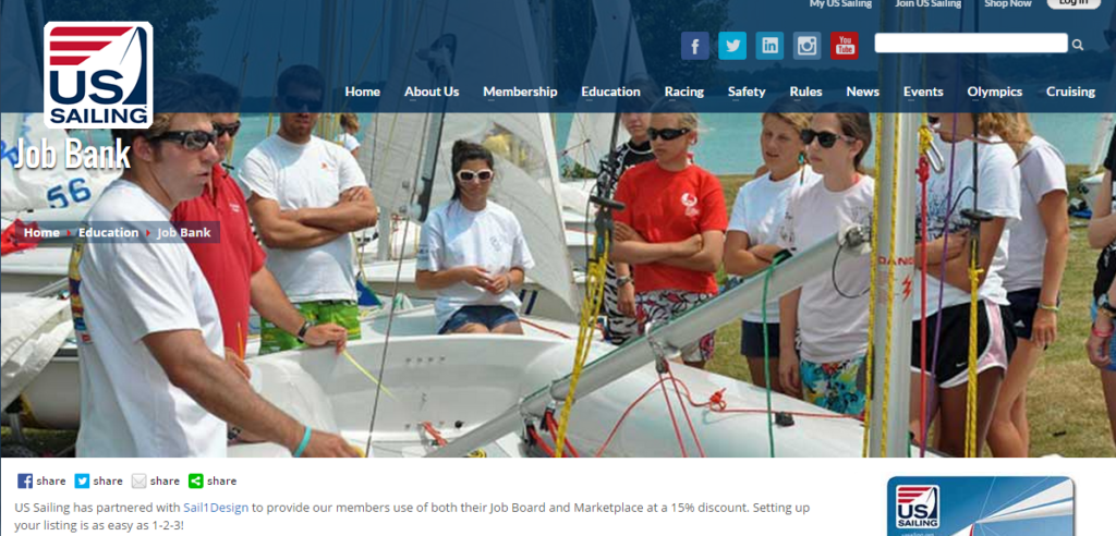 A group of people in sunglasses and casual clothes stand around white sailboats, listening to an instructor. US Sailing logo and website menu are visible at the top. A banner mentions a partnership with Sail1Design.