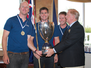 Three men in blue shirts with medals smile as they receive a large silver trophy from an older man in a blazer. Flags are visible in the background.