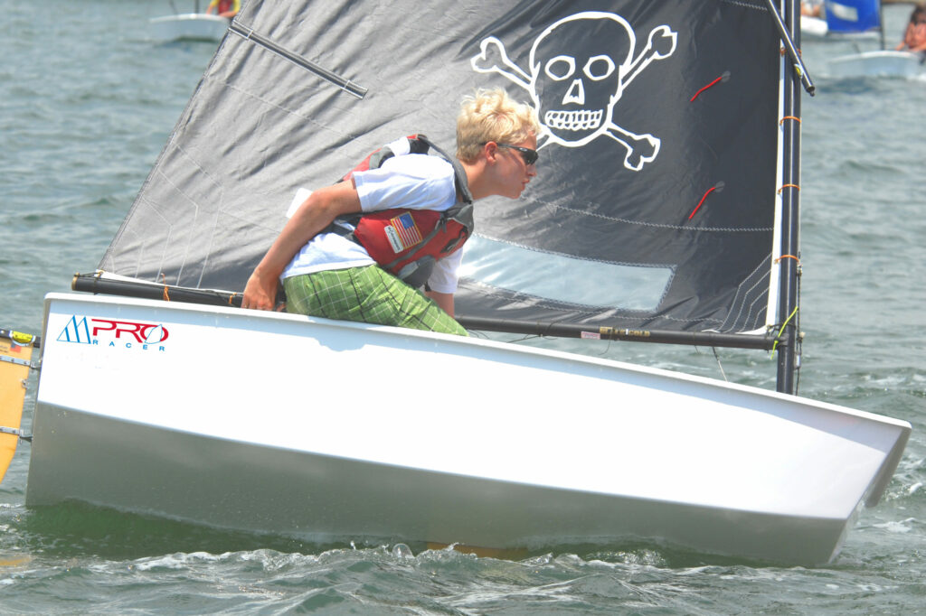 A person with blond hair, wearing sunglasses, a life jacket, and green shorts, sails a small white boat with a black sail featuring a skull and crossbones on a sunny day.