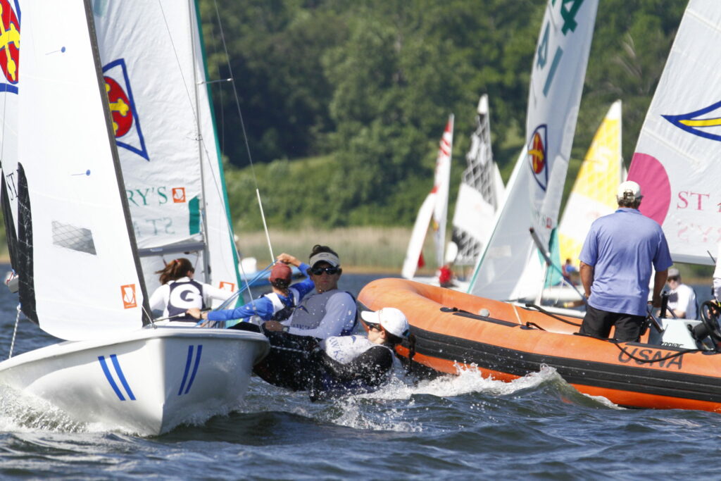 Sailboats and a motorized orange boat with several people on the water during a sailing competition, with trees in the background and sailors leaning out over the edge of their boats.