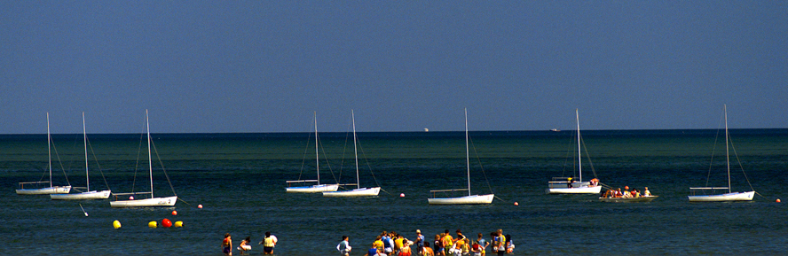 Several white sailboats are anchored in calm blue-green water near the shore, while groups of people gather and swim in the shallow area under a clear sky.