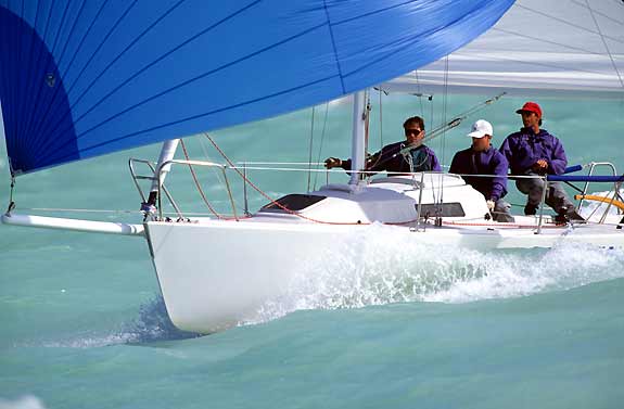 Three people sail a white boat with a large blue sail quickly across turquoise water, creating splashes at the bow.