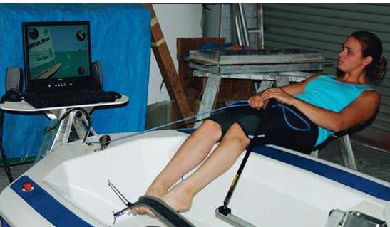 A woman sits in a small boat simulator indoors, holding ropes as she faces a computer monitor displaying a sailing simulation game.