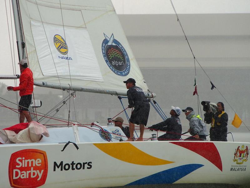 Four people are sailing a yacht in rainy weather. The boat has 'Sime Darby Motors' and 'Malaysia' logos, and the crew members wear rain jackets and caps as they handle the sails and equipment.
