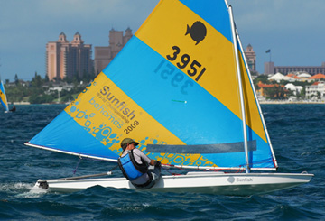 A person wearing a hat and life jacket sails a small white and blue Sunfish sailboat with a colorful yellow and blue sail marked '3951' on the ocean, with city buildings visible in the background.