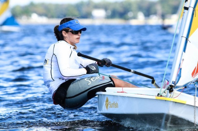 A woman wearing sunglasses and a visor leans out of a small sailboat while gripping the tiller and mainsheet, sailing on a blue body of water with trees in the background.