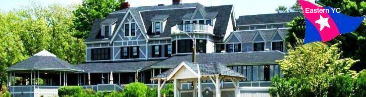 A large, gray shingle-style building with white trim surrounded by trees and greenery; in the upper right corner is a pink, blue, and white flag labeled 'Eastern YC.