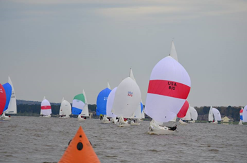 Sailboats with colorful spinnakers race on a lake under a cloudy sky. An orange buoy floats in the foreground, and trees line the distant shore.
