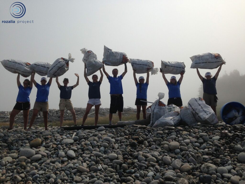 Eight people stand on a rocky beach holding large bags over their heads and smiling. More filled bags and a blue container rest on the ground beside them. Fog and trees are visible in the background.