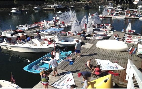 Children and adults prepare small sailboats and dinghies at a busy marina dock on a sunny day, surrounded by water, motorboats, and sailboats. Some people are wearing life jackets and rigging sails.