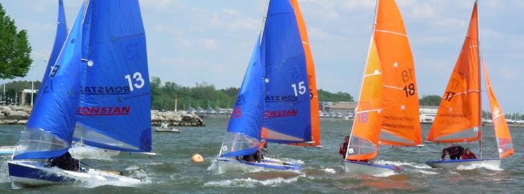Four small sailboats, two with blue sails and two with orange sails, race on a choppy lake near a rocky shoreline and trees under a partly cloudy sky.