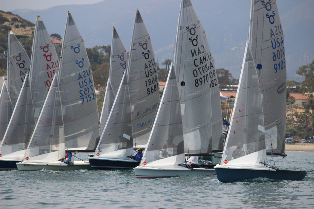 Several sailboats with raised sails, marked with 'USA' and 'CAN,' race closely together on calm water near a shoreline with trees, houses, and distant mountains in the background.