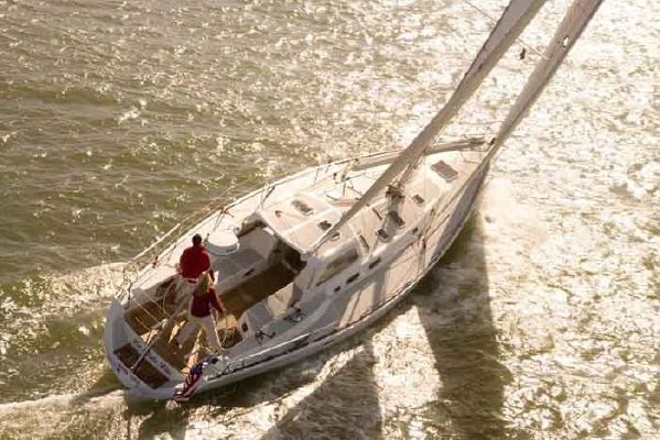 A white sailboat with two people on board sails on sunlit water, leaving a wake behind. The sails are up, and the boat appears to be moving swiftly.