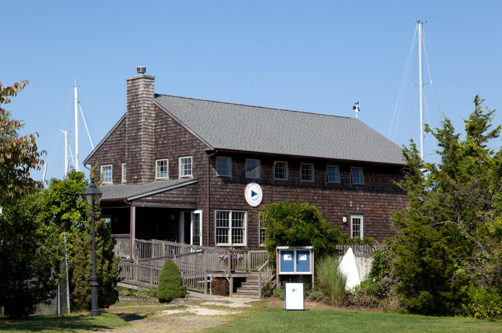A rustic, wooden-sided building with a sloped roof and a large porch sits among greenery, with sailboat masts visible behind it against a clear blue sky. A sign and informational kiosk stand in front.