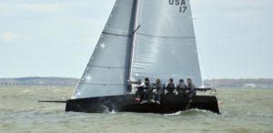 A group of six people sail on a sleek, black sailboat marked 'USA 17' across choppy water, with land and buildings visible in the distant background under a cloudy sky.