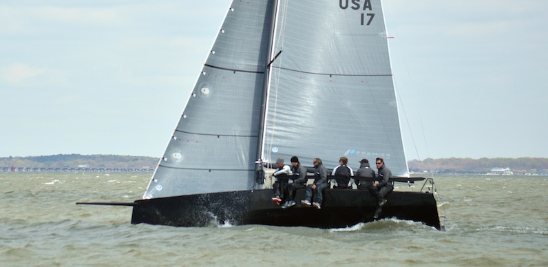 A black sailboat with five people in life jackets sails across choppy water. The crew sits on the edge of the boat, leaning out. The sail has “USA 17” printed on it, and there’s a distant shoreline in the background.