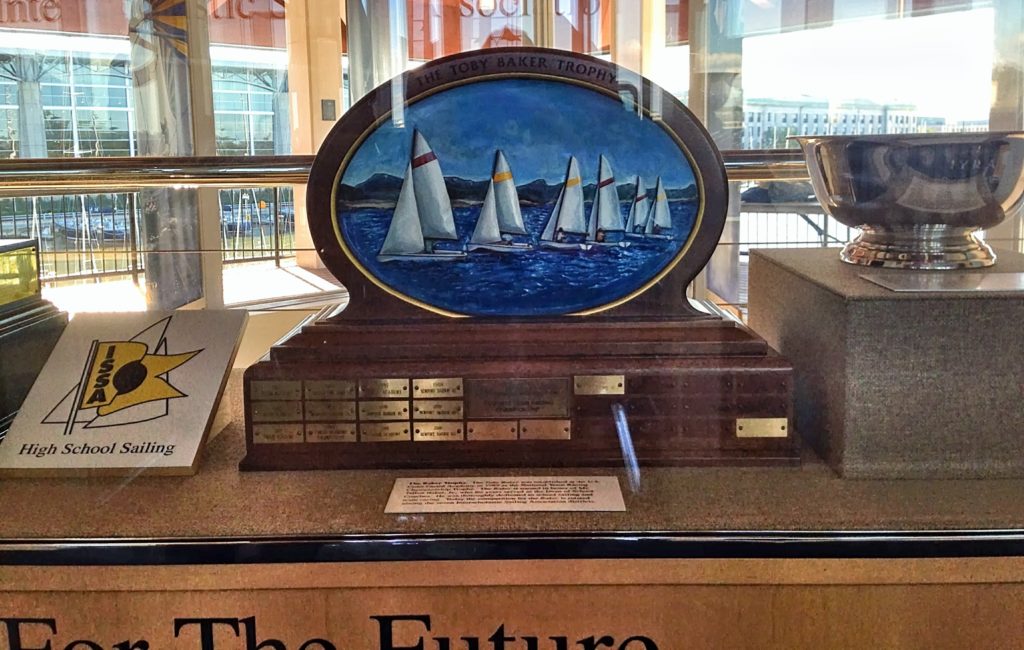 A trophy with a painted oval of sailboats on water, labeled “The Toby Baker Trophy,” displayed in a glass case with plaques, a silver bowl, and a High School Sailing sign nearby.