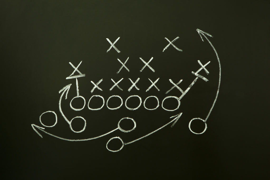 A football play diagram drawn in white chalk on a blackboard, showing Xs and Os representing players with arrows indicating movement and strategy.