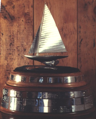 A silver sailboat trophy with a wooden and metal base, featuring engraved plaques, is displayed in front of a wooden wall.