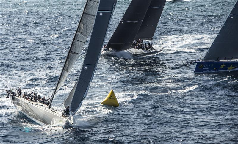 Three sailboats race around a yellow buoy on choppy blue water, their crews actively managing the sails as the boats lean into the wind under a clear sky.