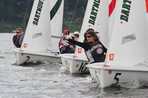 Four sailboats with college teams compete closely on the water. Sailors, wearing sunglasses and team gear, lean out to balance their boats; 'Dartmouth' is visible on sails, and 'Bowdoin' on one sailor's shirt.