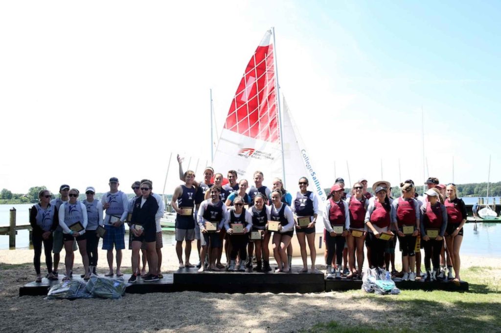 A group of people, many in team uniforms, stand on a dockside platform holding awards in front of a sailboat, with more boats and water in the background on a sunny day.
