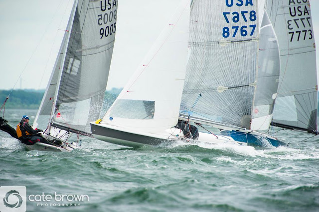 Sailboats racing closely on choppy water, with sailors leaning out to balance the boats. The boat sails display numbers and 'USA.' The scene is energetic under a cloudy sky. 'Cate Brown Photography' watermark is visible.