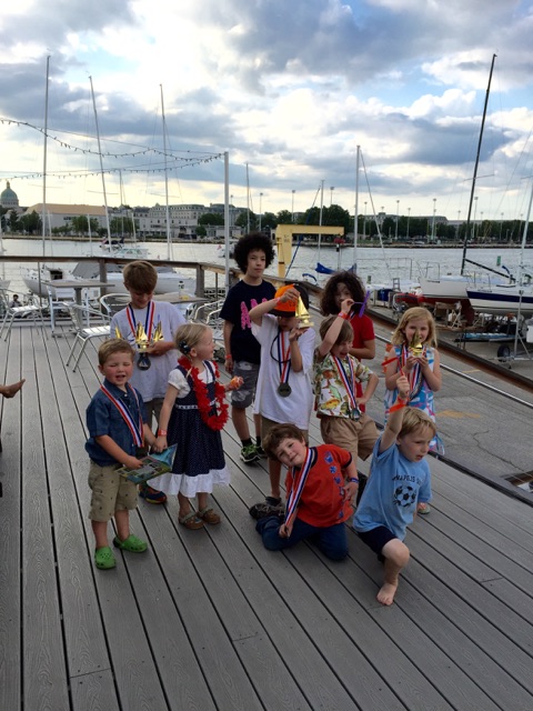 A group of children wearing medals and dress-up accessories stand and kneel on a dock by sailboats, smiling and posing for a photo under a partly cloudy sky.