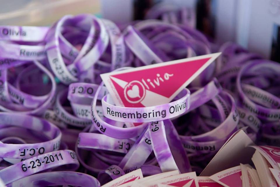 A pile of purple and white marbled wristbands reading 'Remembering Olivia' surrounds a pink heart-shaped sign with 'Olivia' written on it. The wristbands include dates such as '6-23-2011.