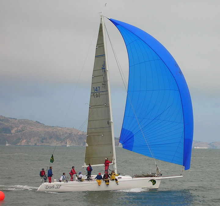 A white sailboat with a large blue spinnaker sail glides on the water. Several people are on board, and hills are visible in the background under a cloudy sky.