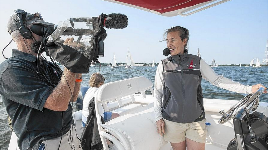 A person smiles and speaks into a headset while steering a boat, being filmed by a cameraman. Sailboats are visible on the water in the background under a clear sky.