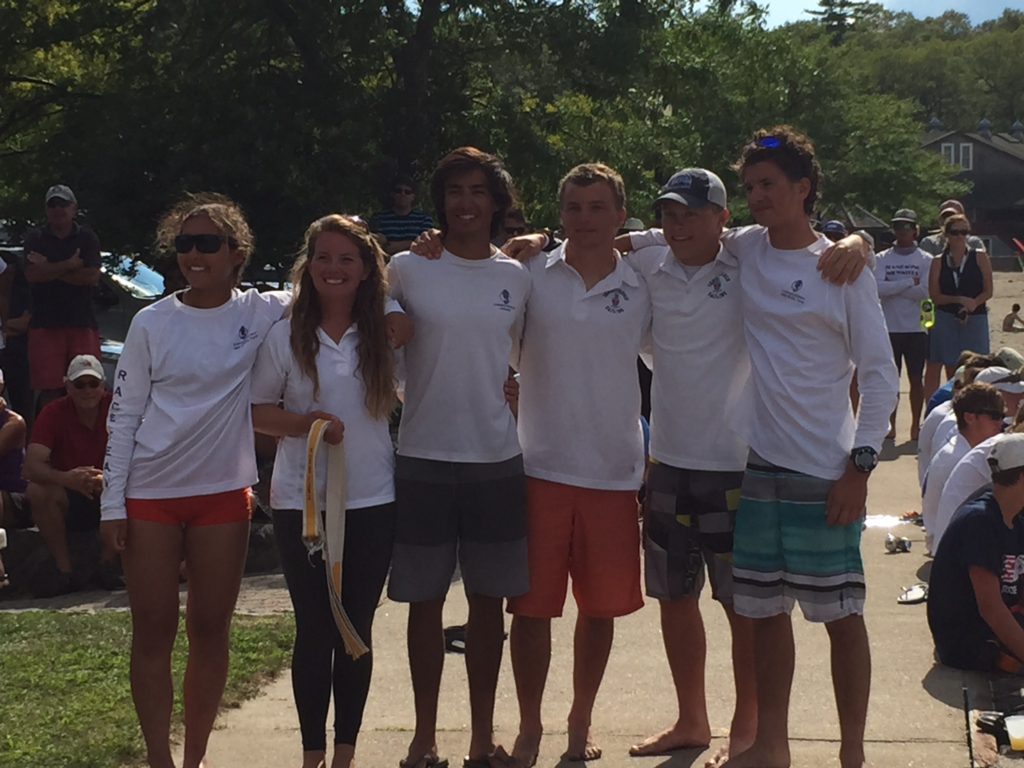 Six young people stand arm-in-arm outdoors, smiling at the camera. They wear matching white long-sleeve shirts and shorts. A crowd sits and stands in the background amid trees and sunlight.