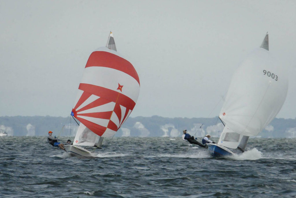 Two small sailboats racing on choppy water, each with two sailors leaning out to balance. The left boat has a red and white sail, while the right boat has a plain white sail. Houses line the distant shore under a cloudy sky.