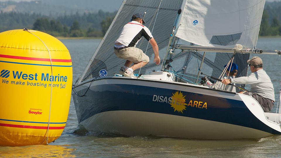 Two people sailing a small boat with 'DISASTER AREA' printed on its side, closely approaching a large yellow West Marine buoy on a lake with trees and hills in the background.