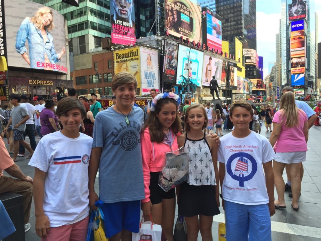 Five smiling teenagers stand together in a busy, colorful Times Square, New York City, surrounded by crowds, billboards, and bright advertisements on a sunny day.