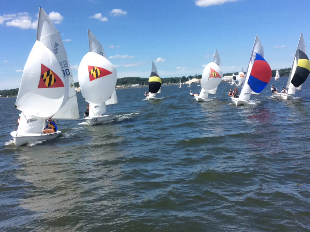 Several small sailboats with colorful spinnakers, including some with Maryland flag designs, race on a lake under a blue sky with scattered clouds. Sailors are visible on each boat as they move through the water.