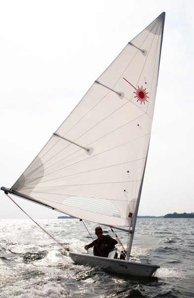 A person sailing a small, single-handed sailboat with a tall, white sail on a sunny day, with waves and distant shoreline visible in the background.