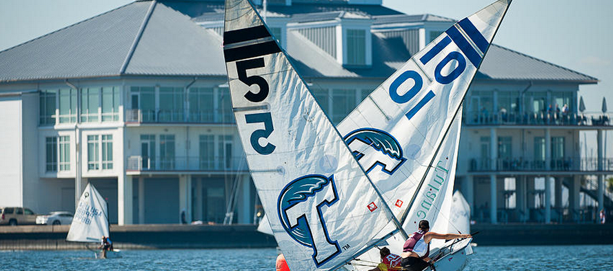 Two small sailboats with the numbers 5 and 10 on their sails race on a body of water in front of a large, white waterfront building with balconies and a gray roof.