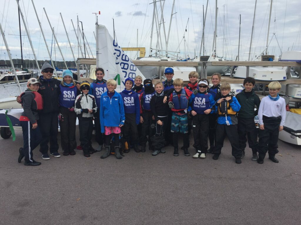 A group of children and adults wearing sailing gear stand together, smiling, in front of sailboats at a marina. Most are wearing jackets with 'SSA' printed on them. The sky is cloudy and many masts are visible in the background.