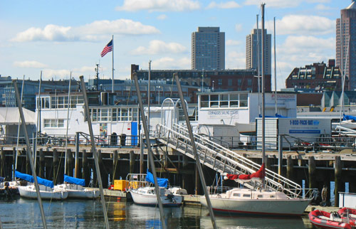 Sailboats docked at a marina with an American flag, white buildings, and tall city buildings in the background under a partly cloudy sky.
