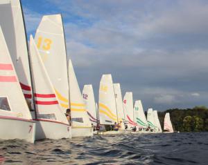 A row of small sailboats with colorful numbered sails lines up on the water, ready to race under a partly cloudy sky with trees in the background.