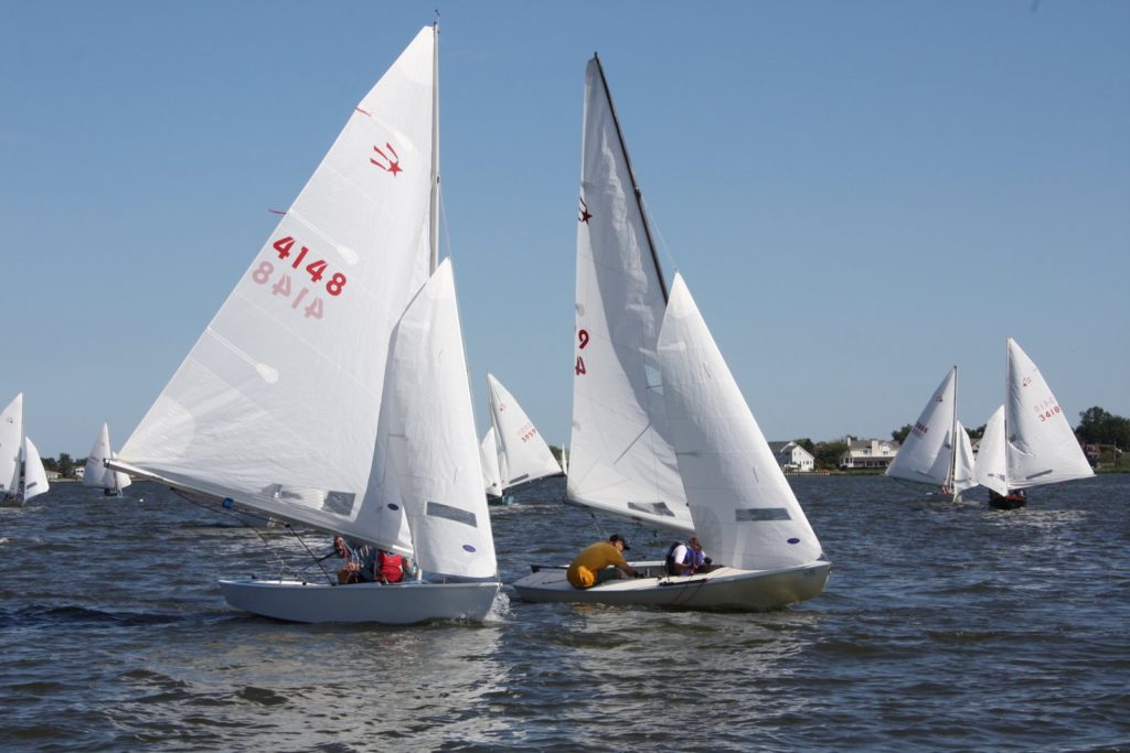 Several small sailboats with white sails are racing on a body of water under a clear blue sky. Some houses and trees are visible on the distant shoreline in the background.