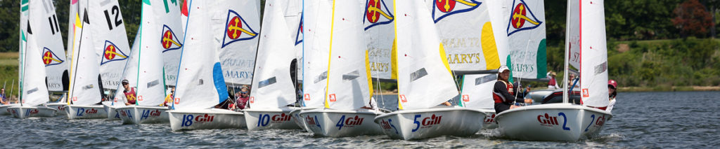 A row of small sailboats with white sails, each marked with colorful logos and numbers, race closely together on a lake with green trees and foliage in the background.
