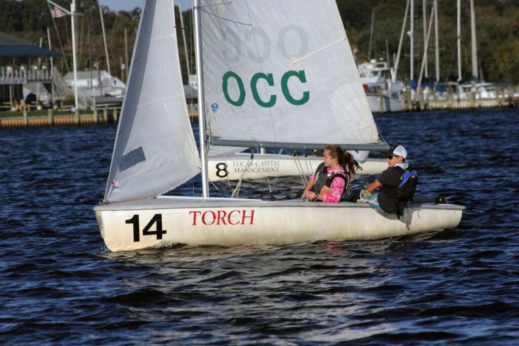 Two people sail a small white boat labeled '14 TORCH' on a body of water. The sail displays 'OCC' and '8.' Trees, boats, and buildings line the distant shore under a clear sky.